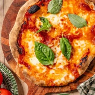 An overhead photo of a neapolitan pizza topped with basil leaves on a wooden platter. Next to this is a plate of tomatoes, a kitchen towel, and a bowl of pizza sauce.