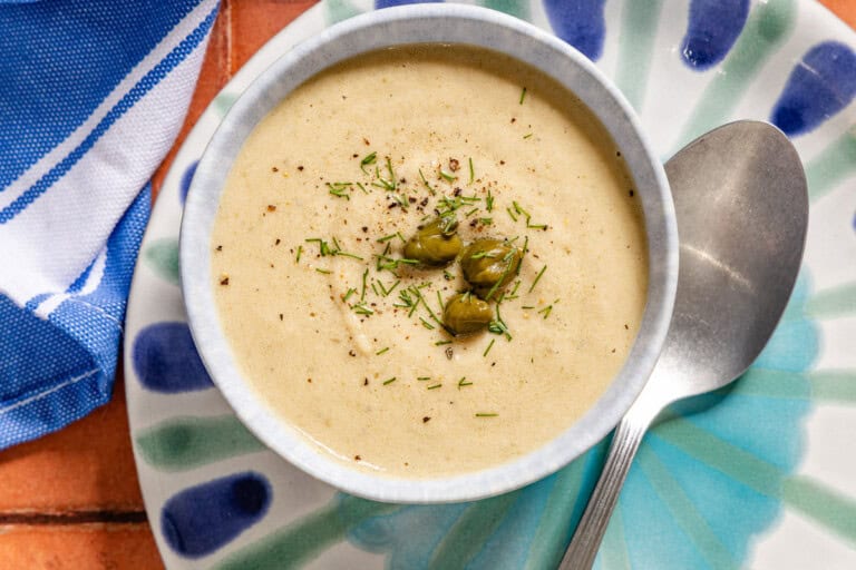 An overhead photo of a bowl of tonnato sauce topped with dill and capers on a plate with a spoon.