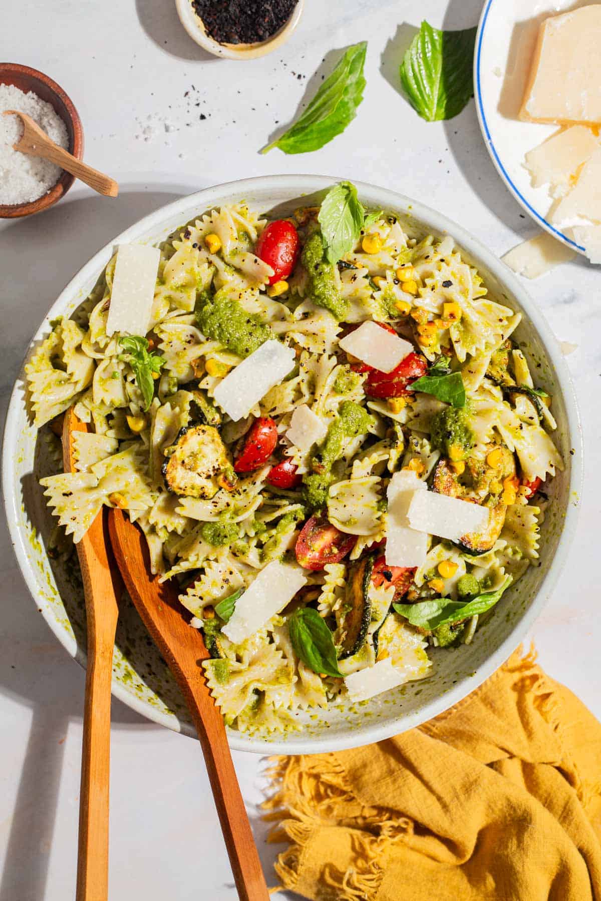An overhead photo of summer pasta salad in a serving bowl with wooden serving utensils. Next to this is a bowl of urfa biber, a bowl of salt, a plate of parmesan cheese and a cloth napkin.