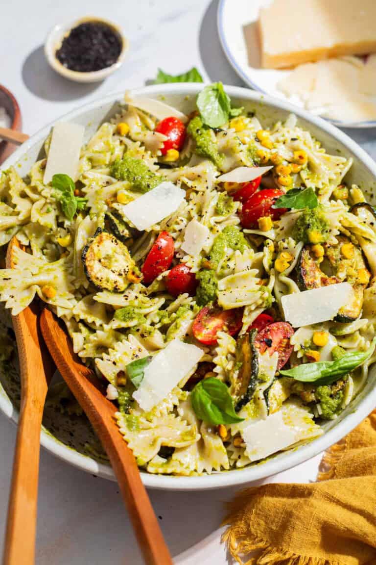 A close up of summer pasta salad in a serving bowl with wooden serving utensils. Next to this is a bowl of urfa biber, a plate of parmesan cheese and a cloth napkin.