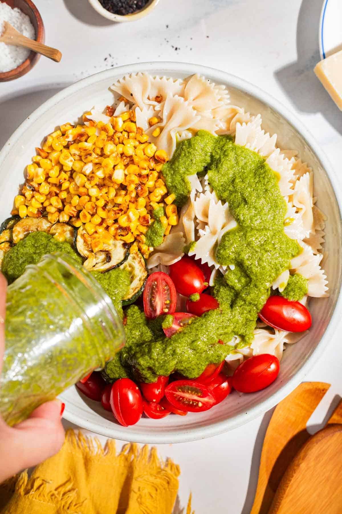 An overhead photo of the basil vinaigrette being poured onto the unmixed summer pasta salad in a bowl. Next to this is a cloth napkin, wooden serving utensils, and bowls of salt and urfa biber.