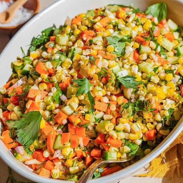 A close up of grilled corn salad in a serving bowl with a spoon. Next to this is a cloth napkin and small bowls of salt and cumin.