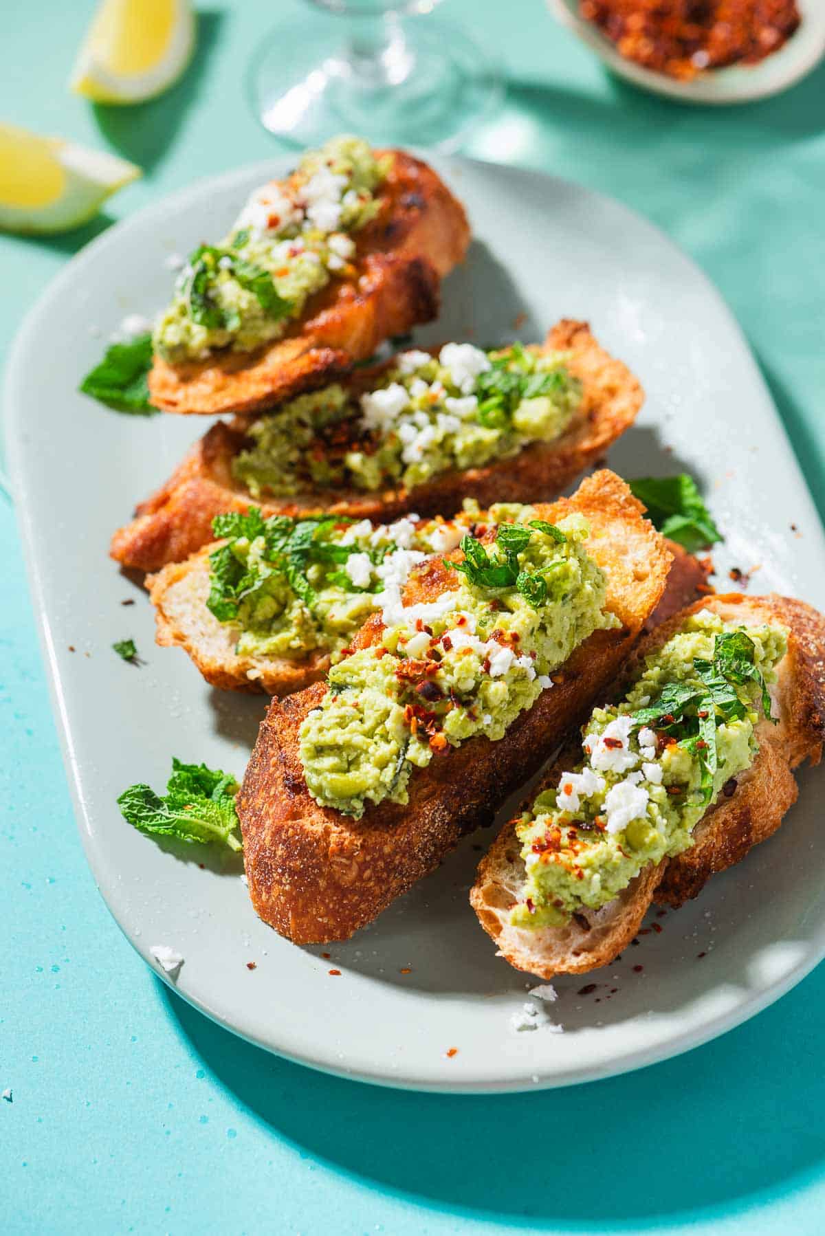 A close up photo of 4 pieces of smashed peas on toast garnished with aleppo pepper, feta and mint on a serving platter. Next to this is a bowl of the smashed peas, a small bowl of Aleppo pepper, and3 pieces of tasted bread.