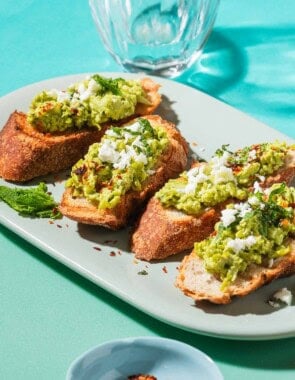 A close up photo of 4 pieces of smashed peas on toast garnished with aleppo pepper, feta and mint on a serving platter. Next to this is a small bowl of Aleppo pepper, and a glass of water.