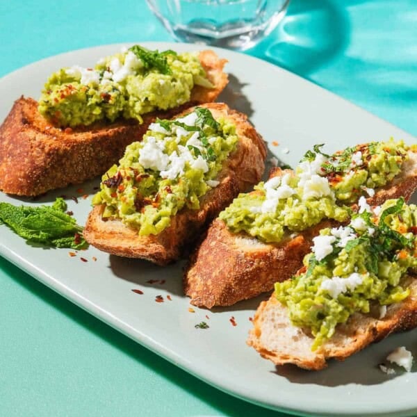 A close up photo of 4 pieces of smashed peas on toast garnished with aleppo pepper, feta and mint on a serving platter. Next to this is a small bowl of Aleppo pepper, and a glass of water.