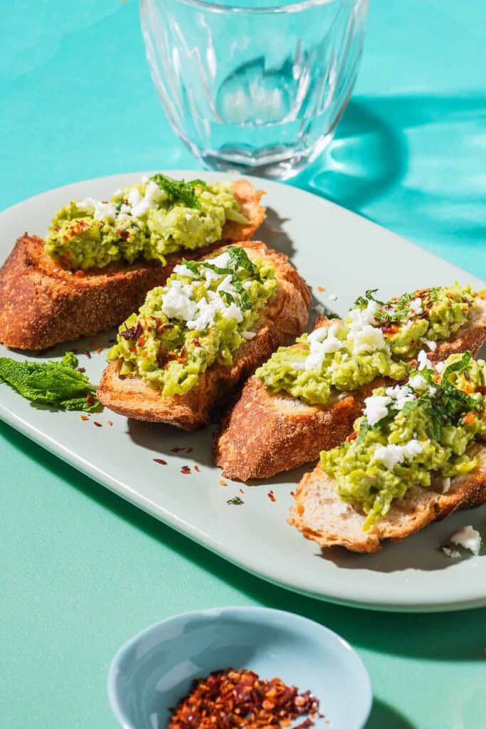 A close up photo of 4 pieces of smashed peas on toast garnished with aleppo pepper, feta and mint on a serving platter. Next to this is a small bowl of Aleppo pepper, and a glass of water.