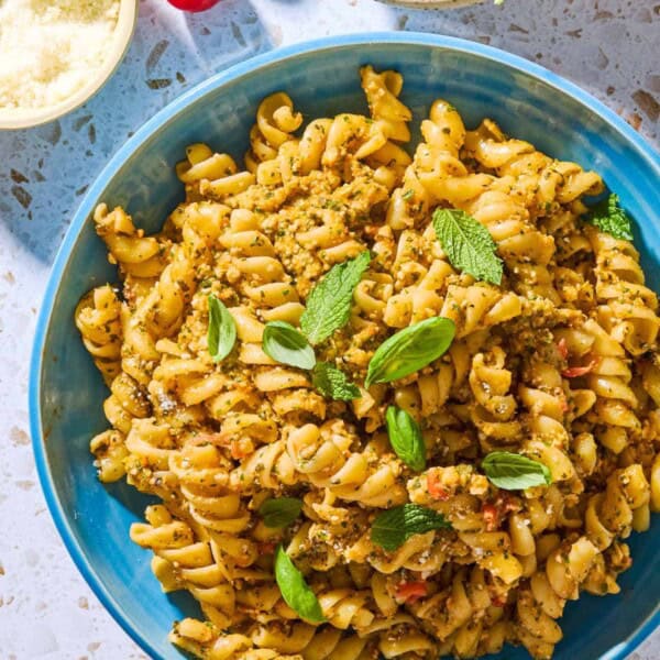 An overhead photo of pesto alla trapanese pasta in a bowl surrounded by 3 cherry tomatoes and bowls of pecorino romano cheese, and basil and mint.