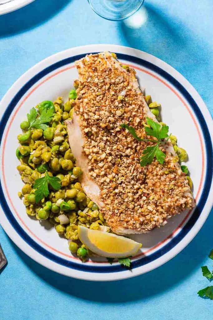 A close up of a serving of baked white fish toppedwith dukkah on a bed of smashed peas on a plate with a lemon wedge.