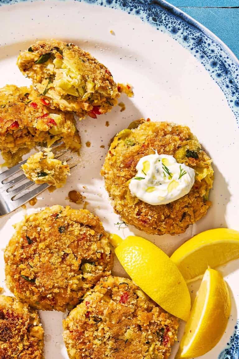 A closeup of vegetarian crab cakes on a serving platter with lemon wedges and a fork. One of the cakes has a small dollop of tzatziki on it.