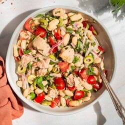 An overhead photo of tuna white bean salad in a serving bowl with a spoon. Next to this is a cloth napkin, sprigs of basil and parsley, and bowls of aleppo pepper and kosher salt.