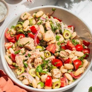 A close up photo of tuna white bean salad in a serving bowl with a spoon. Next to this is a cloth napkin, sprigs of parsley, and bowls of aleppo pepper and kosher salt.