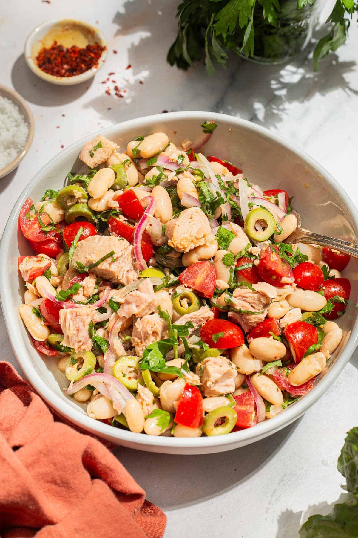 A close up photo of tuna white bean salad in a serving bowl with a spoon. Next to this is a cloth napkin, sprigs of parsley, and bowls of aleppo pepper and kosher salt.