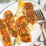 An overhead photo of 4 baked dijon salmon fillets on a serving platter with lemon wedges and a fork. Next to this is a kitchen towel and a plate of lemon wedges.