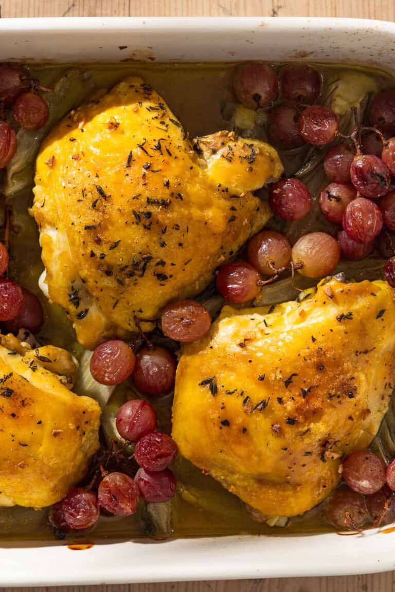 An overhead photo of 3 braised chicken thighs in a baking dish with grapes and fennel.
