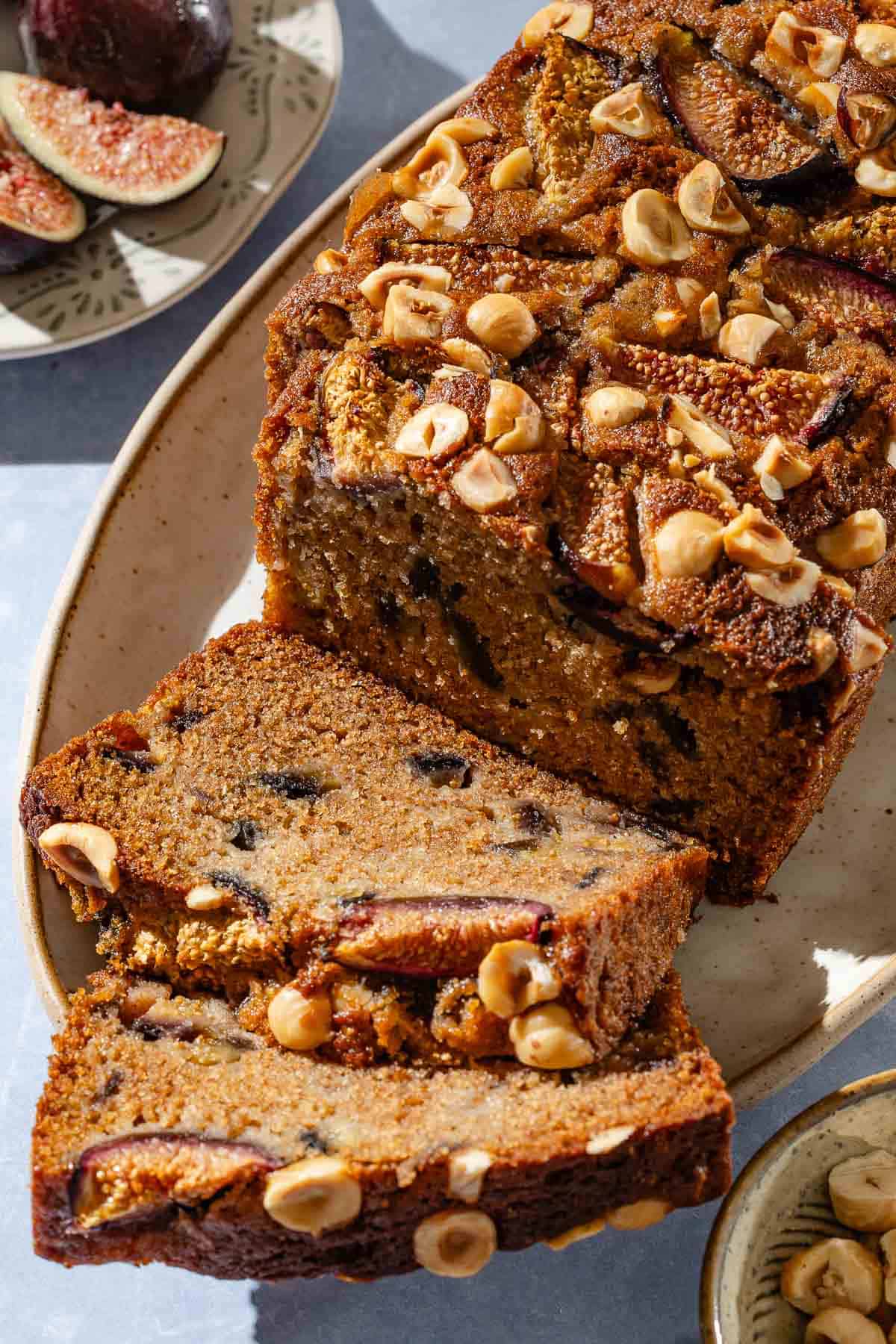 A loaf of partially sliced whole wheat fig bread on a serving plate. Next to this are bowls of hazelnuts and figs.