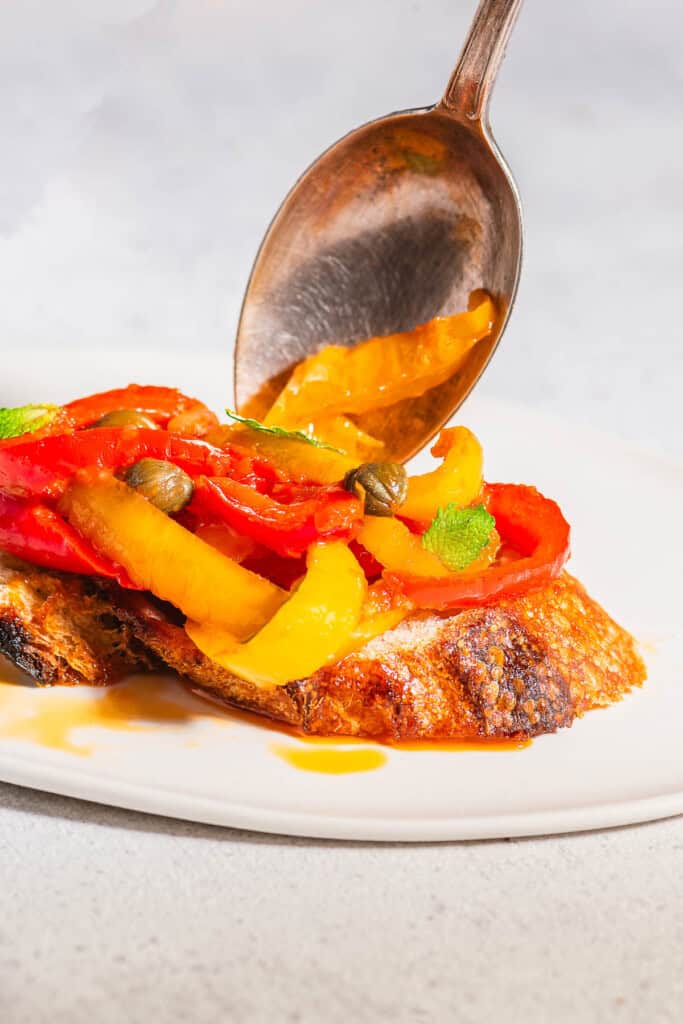 A close up of a sliced grilled bread being topped with peperonata on a plate.