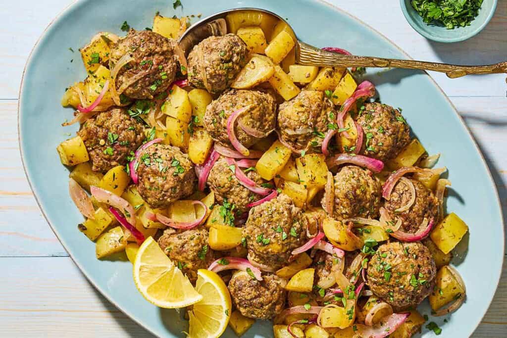 Baked greek meatballs and potatoes on a serving plate with a serving spoon and lemon wedges. Next to this is a small bowl of chopped parsley.