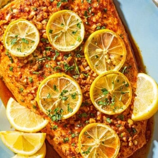 An overhead photo of a lemon garlic salmon fillet topped with lemon wheels and parsley on a serving platter with lemon wedges.