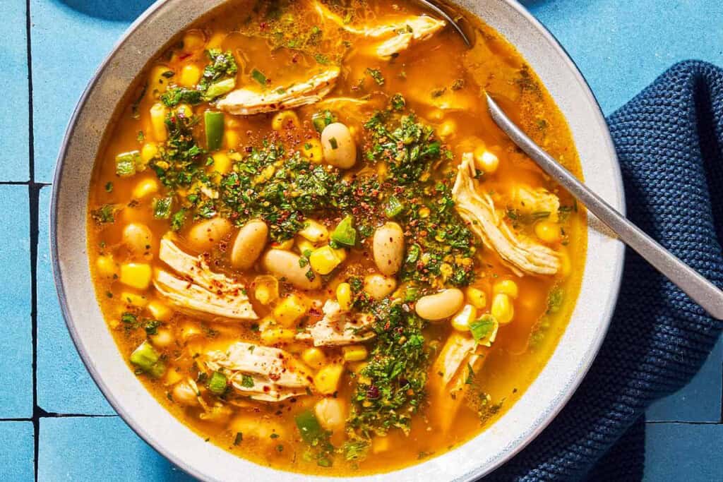 An overhead photo of white chicken chili topped with chermoula in a bowl with a spoon. Next to this is a kitchen towel.