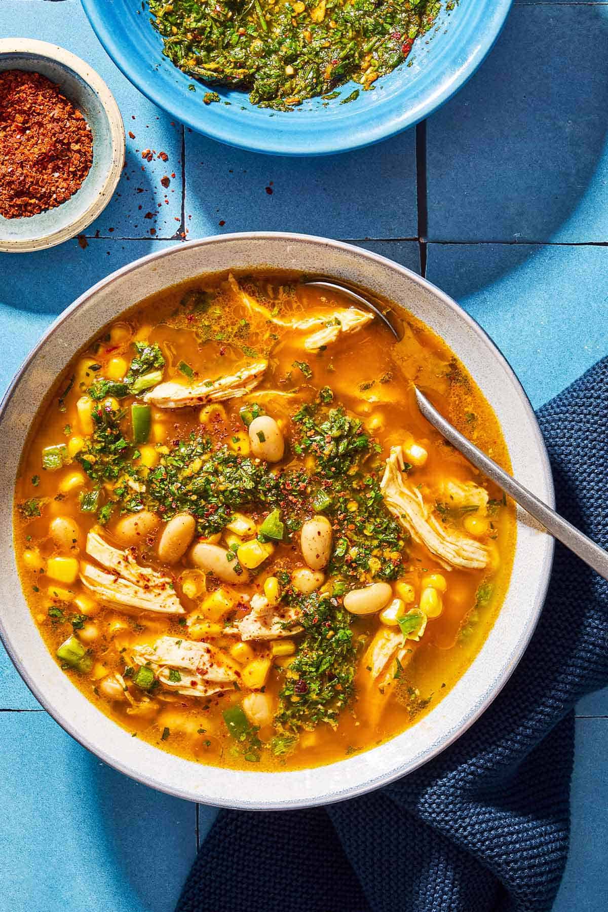An overhead photo of white chicken chili topped with chermoula in a bowl with a spoon. Next to this is a kitchen towel, a bowl of aleppo pepper and a bowl of chermoula.