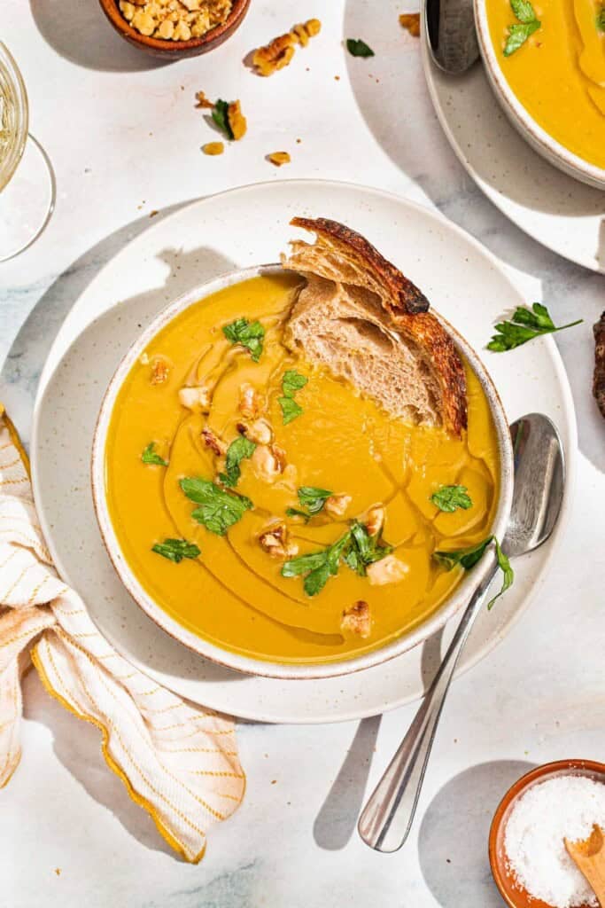 An overhead photo of acorn squash soup garnished with walnuts, parsley and a drizzle of olive oil in a bowl with a piece of bread. Next to this is a spoon, a cloth napkin, another bowl of the soup, and small bowls of walnuts and salt.