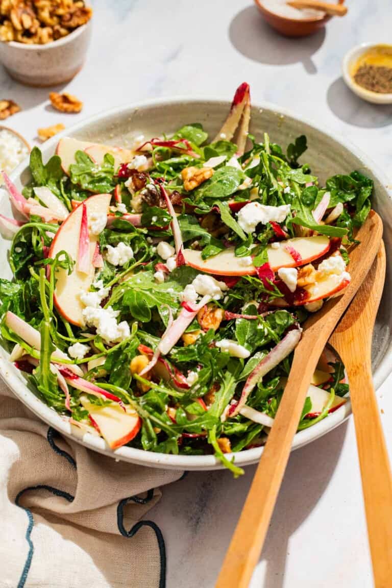 Apple walnut salad in a serving bowl with wooden serving utensils surrounded by bowls of salt and pepper and a cloth napkin.