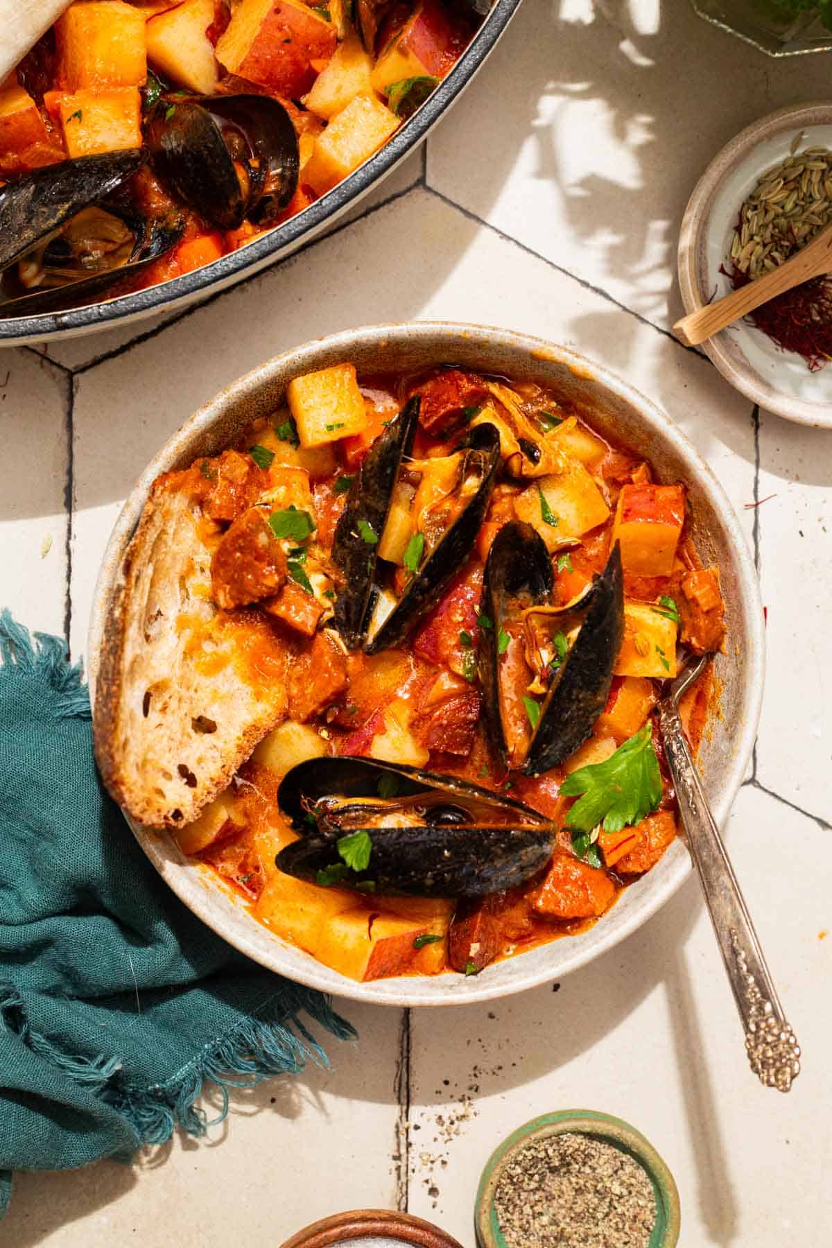 An overhead photo of a bowl of mussels with chorizo in a saffron stew with a fork. Next to this is the pot with the rest of the stew, a cloth napkin, and bowls of spices.