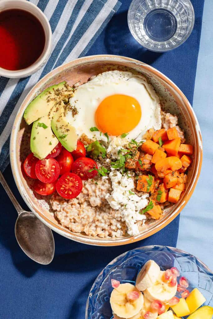 An overhead photo of a savory oatmeal bowl surrounded by a bowl of fruit, a spoon, a glass of water and a cup of tea.