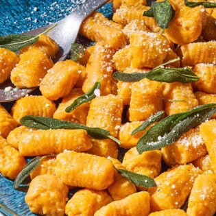A close up of sweet potato gnocchi and sauce topped with fresh sage on a serving platter with a spoon.