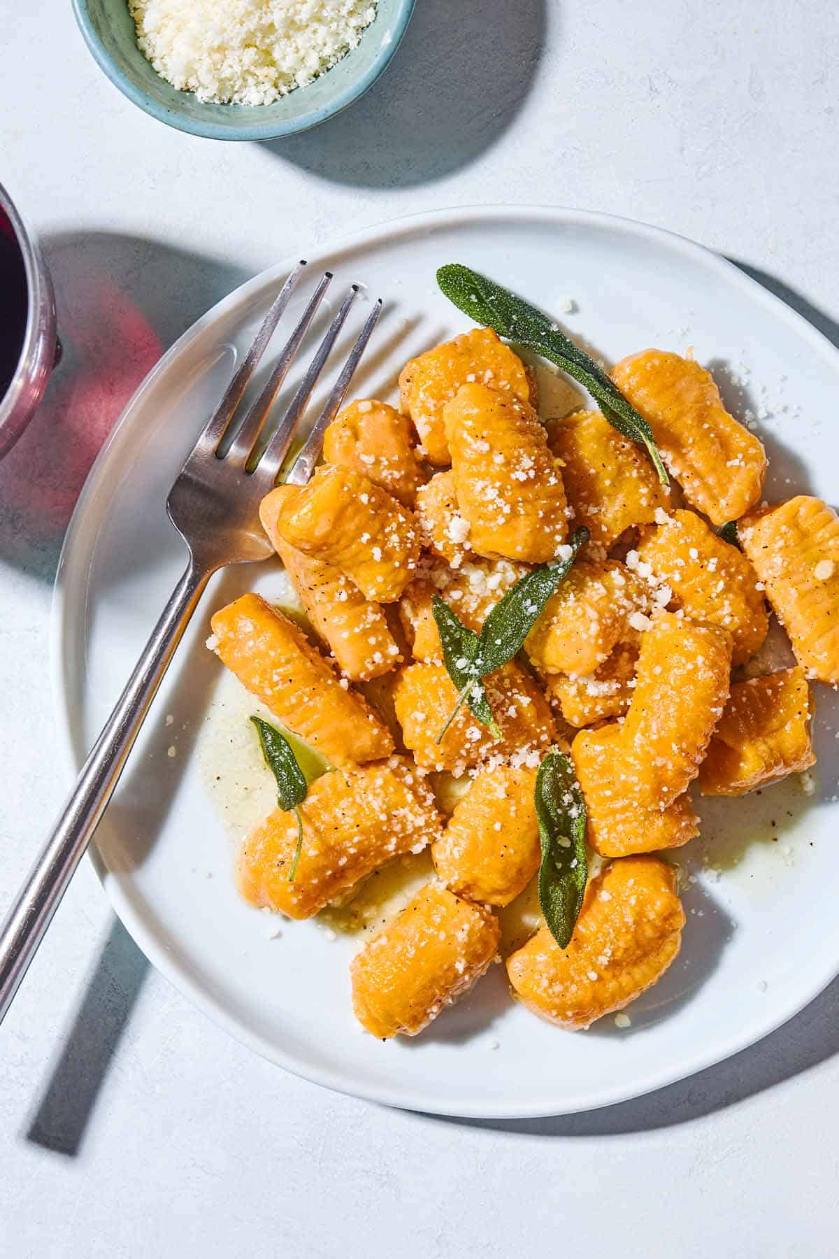A close up of sweet potato gnocchi and sauce topped with fresh sage and parmigiano-reggiano cheese on a plate with a fork. Next to this is a bowl of the grated parmigiano-reggiano cheese.