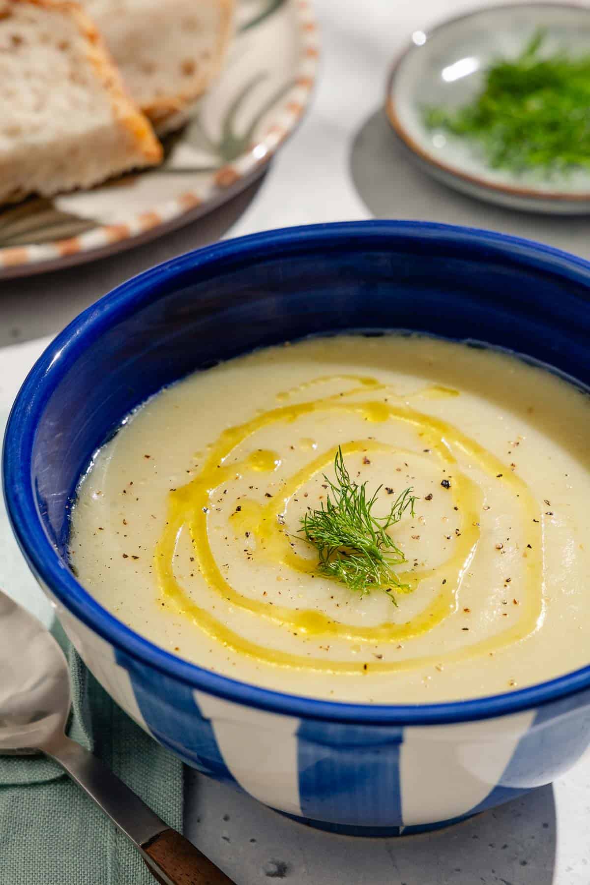 A close up of a bowl of fennel soup topped with a drizzle of olive oil and fennel fronds. Next to this is a cloth napkin, spoon, and plates of sliced bread and fennel fronds.