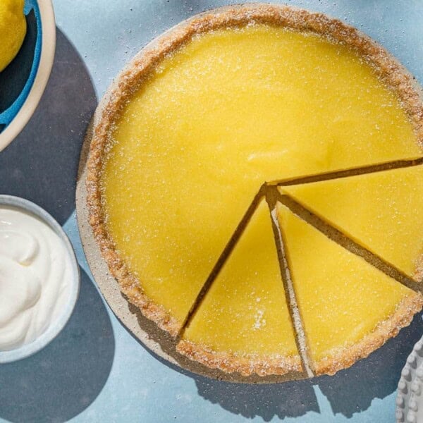 An overhead photo of a lemon tart with 3 pieces sliced. Next to this is a stack of 3 plates with 2 forks, and bowls of lemons, berries and greek yogurt.