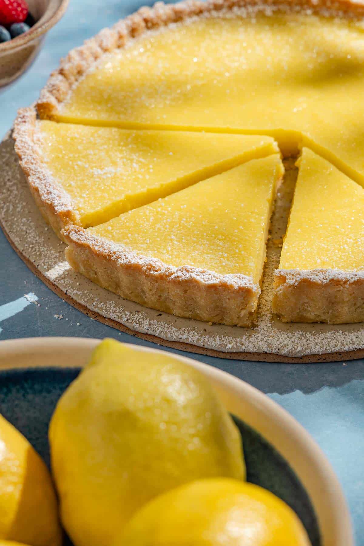 A close up of a lemon tart with 3 pieces sliced next to a bowl of lemons.