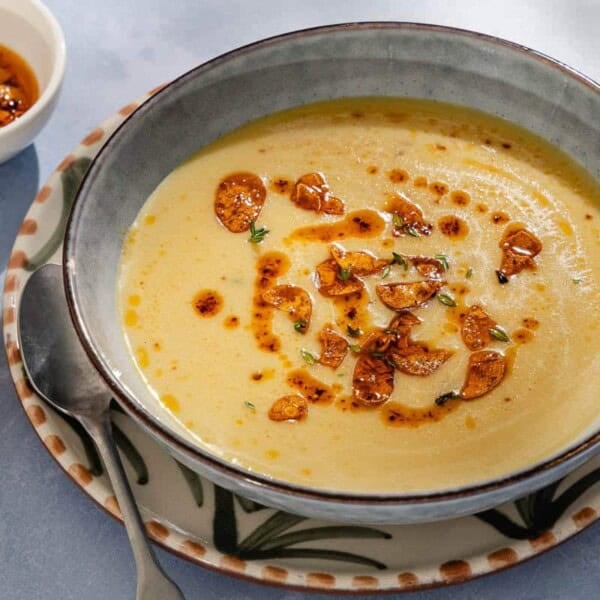 A close up of garlic soup topped with fried garlic and aleppo oil in a bowl on a plate with a spoon. Next to this is a piece of sliced bread and a small bowl of the fried garlic and aleppo oil.