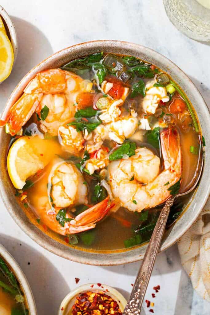 An overhead photo of a bowl of seafood stew with a spoon. Next to this is a glass of white wine and a small bowl of red pepper flakes.