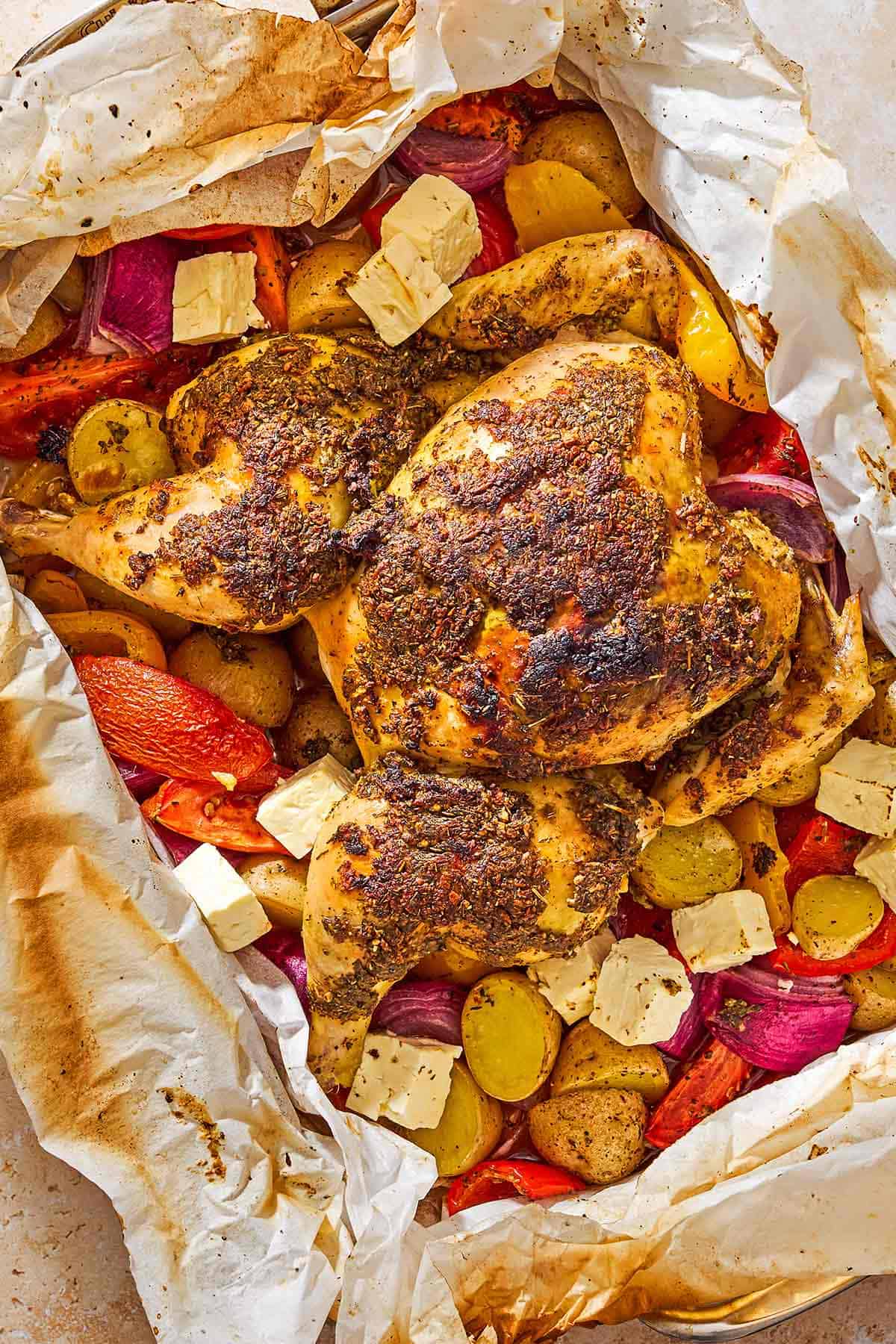 An overhead photo of oven roasted whole chicken and potatoes along with other vegetables and cubed feta in a parchment-line roasting pan.