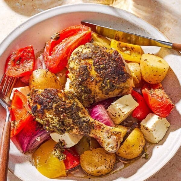 An overhead photo of a serving of oven roasted whole chicken and potatoes along with other vegetables and cubed feta on plate with a knife and fork. Next to this is a cloth napkin, a glass of white wine, and a bowl of Italian seasoning.