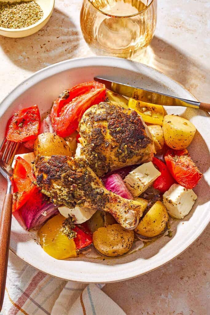 An overhead photo of a serving of oven roasted whole chicken and potatoes along with other vegetables and cubed feta on plate with a knife and fork. Next to this is a cloth napkin, a glass of white wine, and a bowl of Italian seasoning.