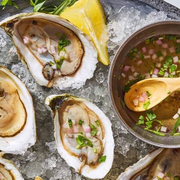 A close up of 3 raw oysters with mignonette sauce on a bed of ice on a tray with lemon wedges and a bowl with the rest of the sauce and a spoon.