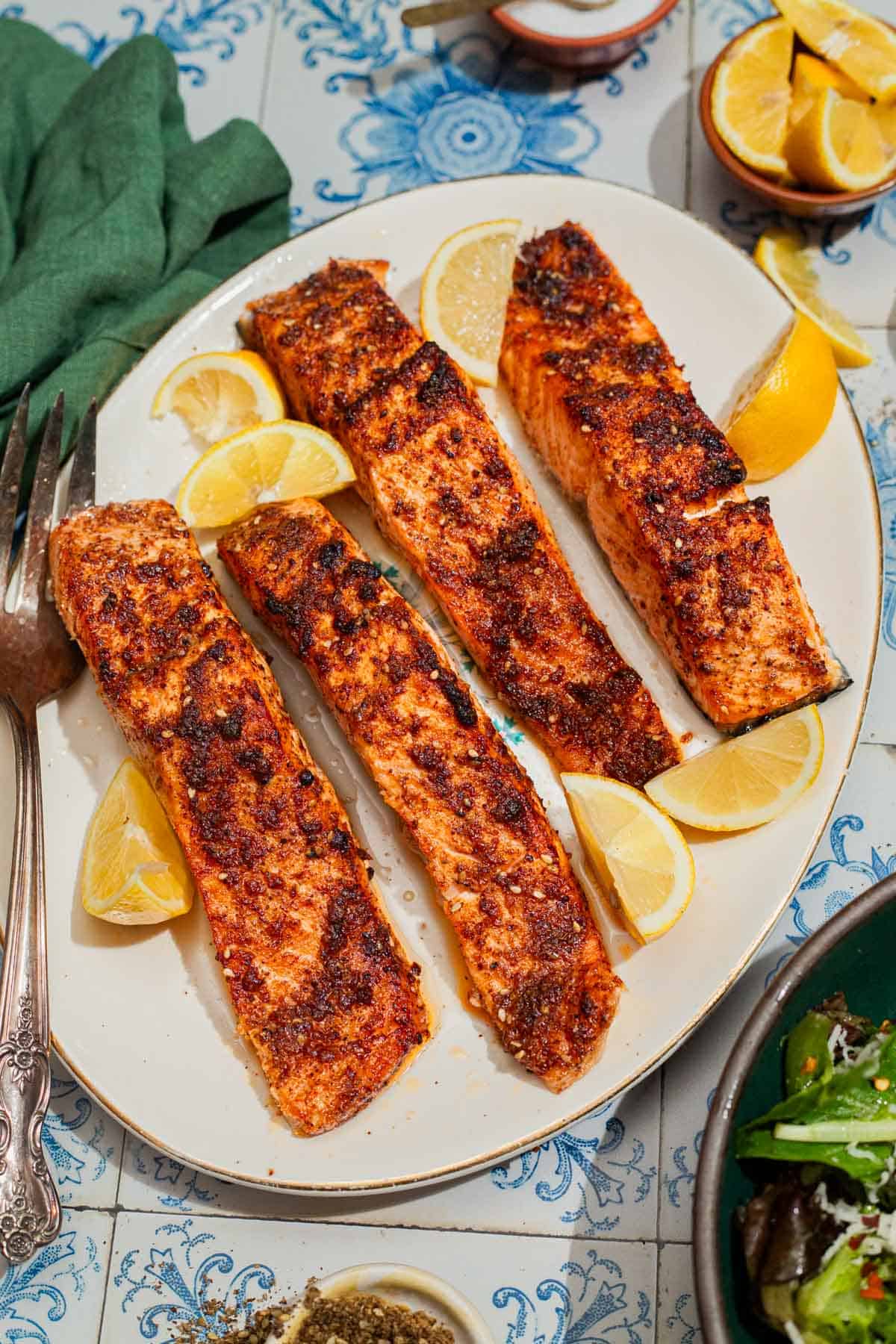 An overhead photo of 4 air fried salmon fillets on a platter with a fork and some lemon wedges. Next to this is a cloth napkin and a bowl of lemon wedges.