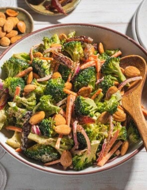 A close up of broccoli salad in a serving bowl with a wooden spoon. Next to this is a stack of 2 plates and small bowls of dates and toasted almonds.