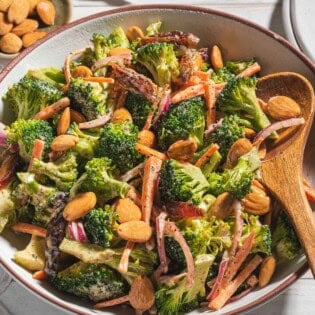 A close up of broccoli salad in a serving bowl with a wooden spoon. Next to this is a stack of 2 plates and small bowls of dates and toasted almonds.
