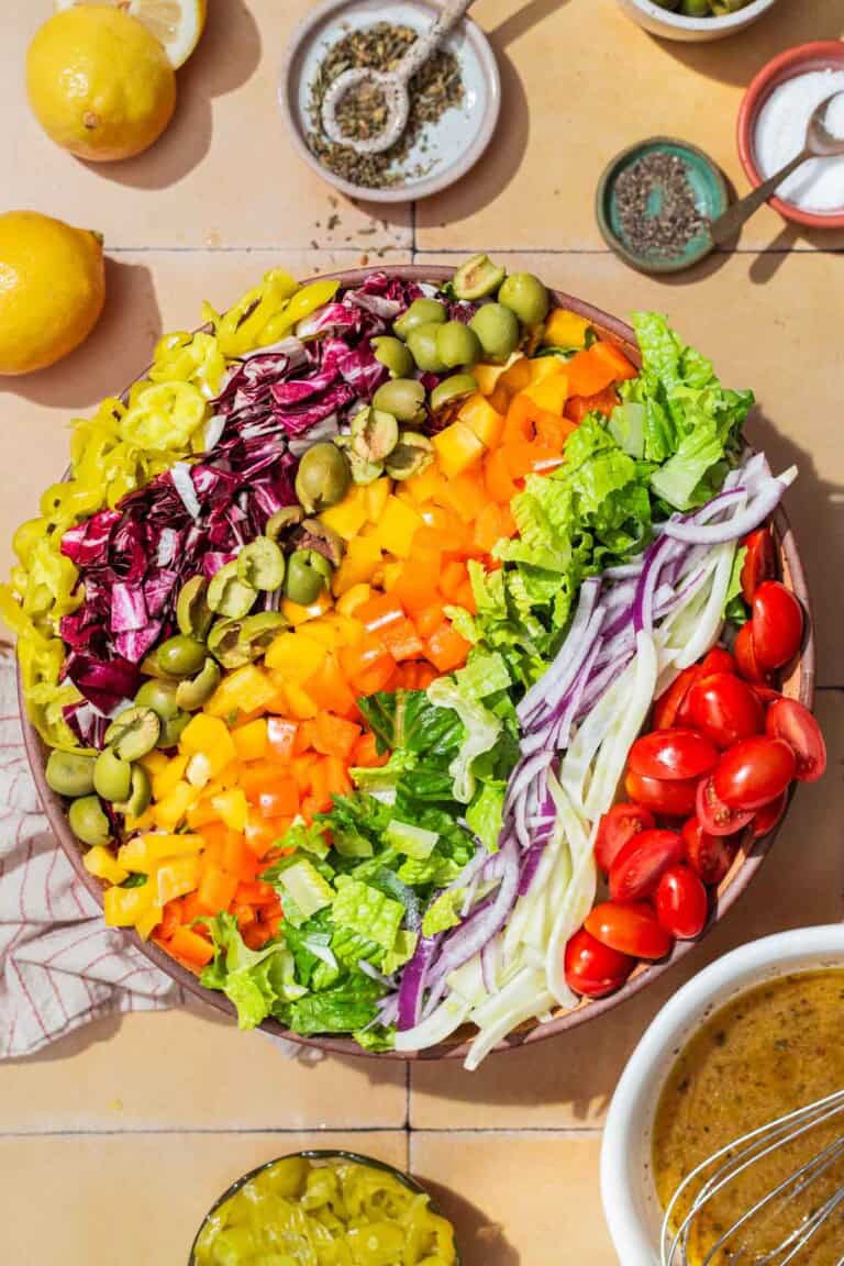 An overhead photo of the ingredients for the chopped salad neatly arranged in a serving bowl, just before being tossed together. Next to this is a bowl of the vinaigrette, lemons, a cloth napkin, and bowls of salt, pepper, and pepperoncini.