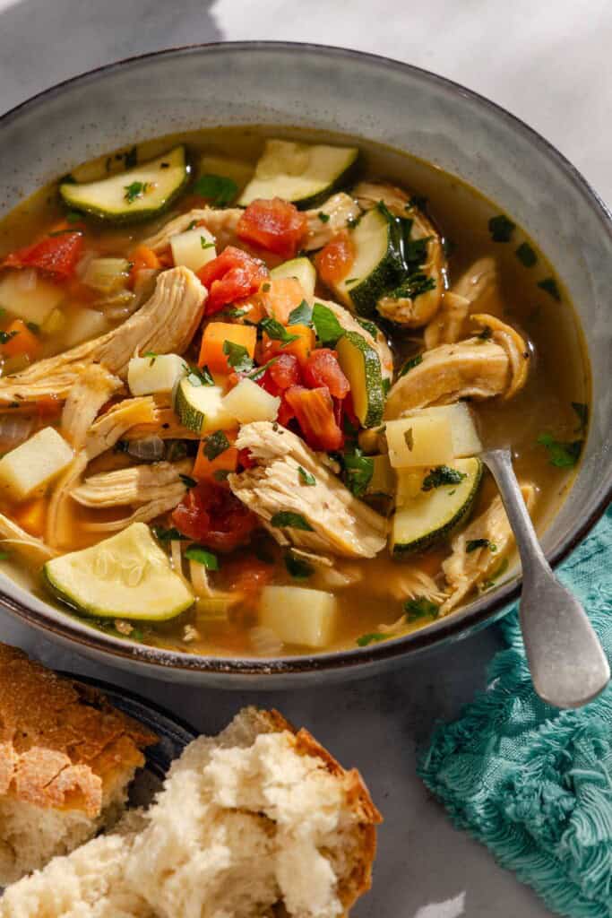 A close up of a serving of chicken vegetable soup in a bowl with a spoon next to pieces of crusty bread.