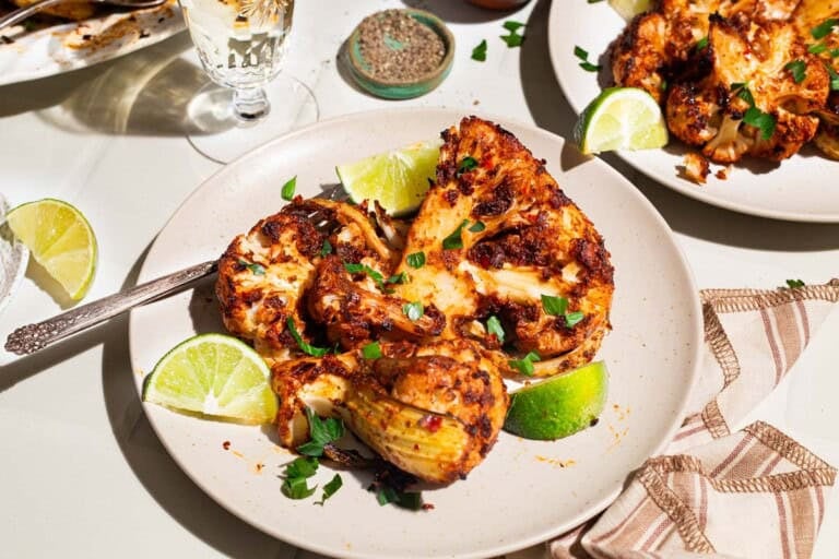 A close up of a serving of spicy baked cauliflower topped with chopped parsley and lime wedges on a plate with a fork. Next to this is a plate with another serving of the cauliflower, a glass of white wine, and a bowl of black pepper and a cloth napkin.