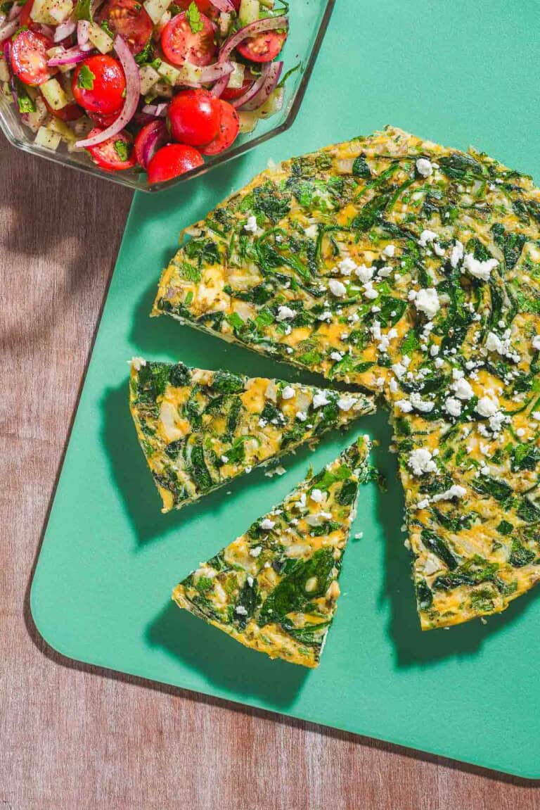 An overhead photo of a feta spinach frittata on a cutting board with two slices cut from it. Next to this is a mediterranean cucumber salad in a bowl.