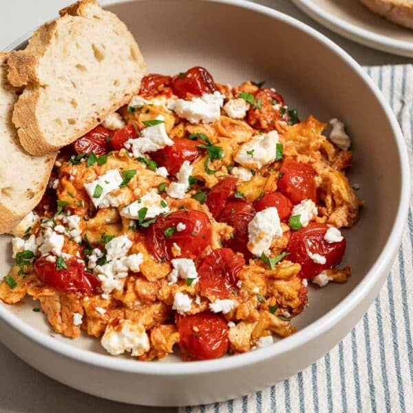 A close up of a serving of strapastada greek tomato scrambled eggs topped with crumbled feta and chopped parsley in a bowl with 2 slices of bread. Next to this is a cloth napkin and a plate of sliced bread.