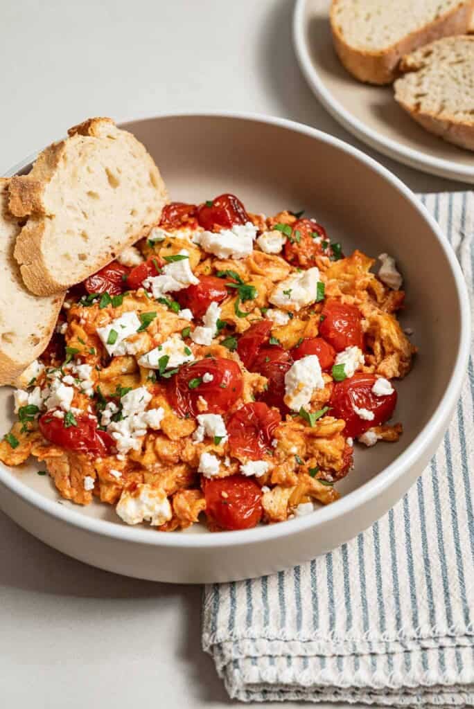 A close up of a serving of strapastada greek tomato scrambled eggs topped with crumbled feta and chopped parsley in a bowl with 2 slices of bread. Next to this is a cloth napkin and a plate of sliced bread.