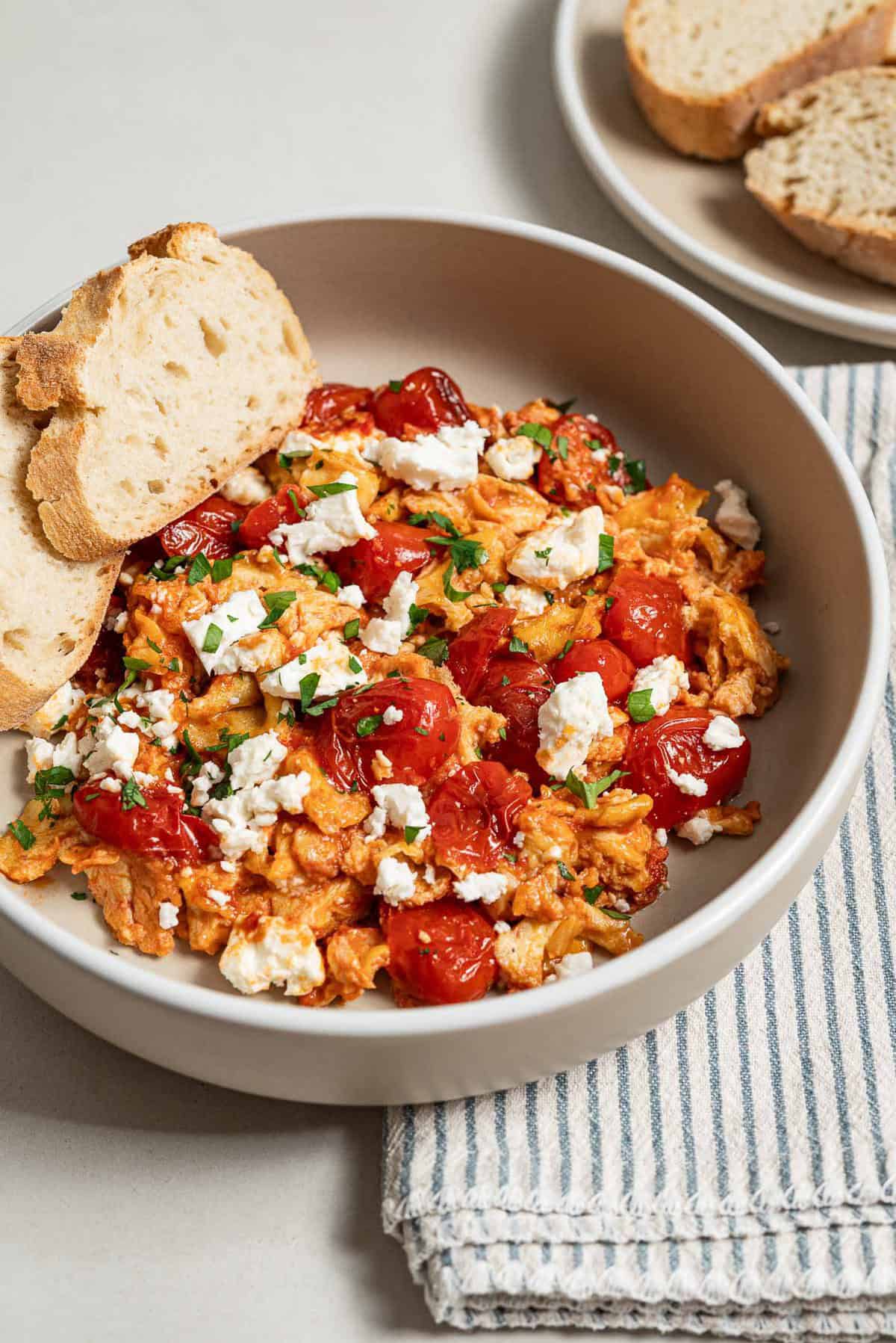 A close up of a serving of strapastada greek tomato scrambled eggs topped with crumbled feta and chopped parsley in a bowl with 2 slices of bread. Next to this is a cloth napkin and a plate of sliced bread.