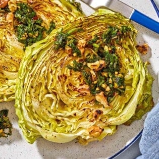 A close up of two cabbage steaks topped with green chermoula and toasted walnuts on a plate with a fork.